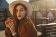 Outdoor fashion portrait of young elegant fashionable brunette woman, model wearing stylish white hat, wrist watch,  brown faux fur coat, posing at sunset, in European city. Copy empty space for text