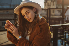 Outdoor Fashion Portrait Of Young Elegant Fashionable Brunette Woman, Model Wearing Stylish White Hat, Wrist Watch,  Brown Faux Fur Coat, Posing At Sunset, In European City. Copy Empty Space For Text