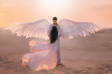 Men Angel Hugs Young Woman. Long Dark Hair Dress Silk Fabric Flying Flutter Wind. Costume Huge White Wings. Bright Color Peach Pink Yellow Sunset Desert. Shoot Back Rear View Turned Away Without Face