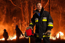 Horizontal View. Firefighter With Sad Equipment, Holds The Helmet In His Hand, Looking Up, Which Stands Against The Background Of A People And Forest In Flames.
