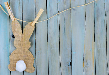 Bunny shape in burlap with a cotton swab tail hanging from twine by clothespins. Background is blue washed rustic wood planks. Copy space.