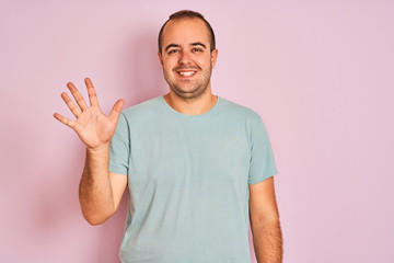 Sticker - Young man wearing blue casual t-shirt standing over isolated pink background showing and pointing up with fingers number five while smiling confident and happy.