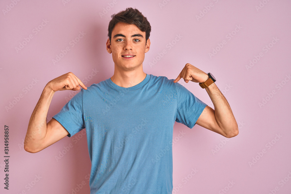Teenager boy wearing casual t-shirt standing over blue isolated background looking confident with smile on face, pointing oneself with fingers proud and happy. - obrazy, fototapety, plakaty 