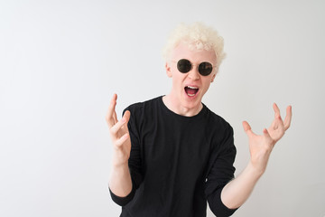 Wall Mural - Young albino man wearing black t-shirt and sunglasess standing over isolated white background crazy and mad shouting and yelling with aggressive expression and arms raised. Frustration concept.