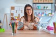© Krakenimages.com - Young beautiful teacher woman wearing sweater and glasses sitting on desk at kindergarten happy face smiling with crossed arms looking at the camera. Positive person.