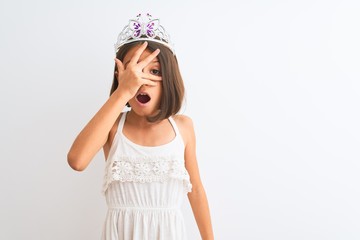 Sticker - Beautiful child girl wearing princess crown standing over isolated white background peeking in shock covering face and eyes with hand, looking through fingers with embarrassed expression.