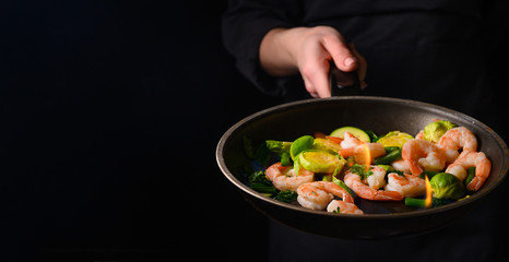 Chef cooks shrimps with vegetables, in a pan with fire. Frying and cooking. Oriental cuisine. Culinary, restaurant business, healthy and tasty food. Banner, on black background.