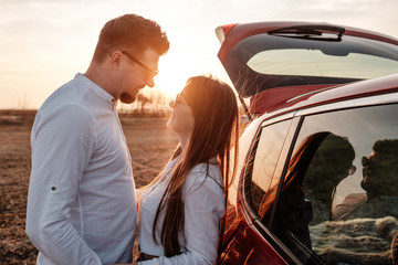 Wall Mural - Young Happy Couple Dressed Alike in White Shirt and Jeans Enjoying Road Trip at Their New Car, Beautiful Sunset on the Field, Vacation and Travel Concept