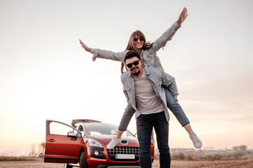 Wall Mural - Young Happy Couple Dressed Alike in White Shirt and Jeans Enjoying Road Trip at Their New Car, Beautiful Sunset on the Field, Vacation and Travel Concept