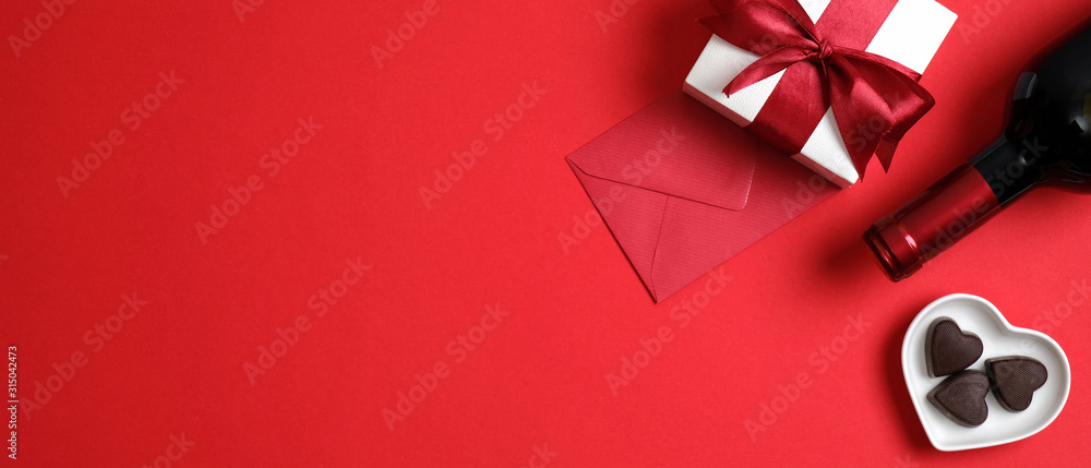 Happy Valentines day banner. Top view table setting for Valentines day with bottle of wine champagne, gift box with red ribbon bow, heart shaped candies and envelope on red background with copy space - obrazy, fototapety, plakaty 