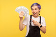 © khosrork - Portrait of surprised stylish hipster girl with violet short hair in denim overalls pointing at fan of dollar banknotes, looking amazed with open mouth. isolated on yellow background, studio shot