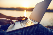 Close Up Of Caucasian Man Sitting In Nature And Using Laptop. In Background Is River And Sunset. Selective Focus On Hands.