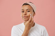 © Seventyfour - Portrait of beautiful young woman applying face cream and looking at camera while posing against pink background during beauty routine in morning
