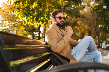 Poster - Image of handsome adult man drinking coffee while sitting on bench in park