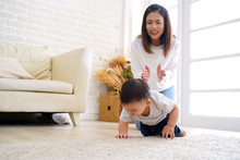 Serious Asian Little Boy Crawling On Floor While Young Mother Sitting Behind Cheering Up And Clapping Hands In Light Living Room