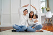 © twinsterphoto - Positive young Asian married couple with infant boy looking up and making roof figure with hands while sitting on carpet in light room