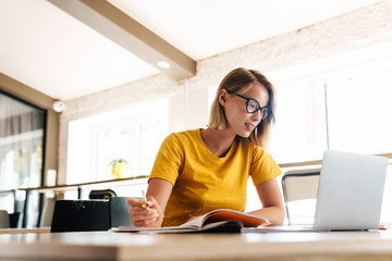 Photo of beautiful young woman making notes and using laptop