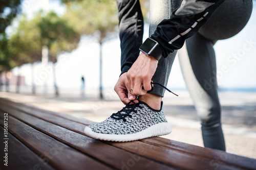 Photographie A young sportswoman with smartwatch outdoors in city, tying shoelaces