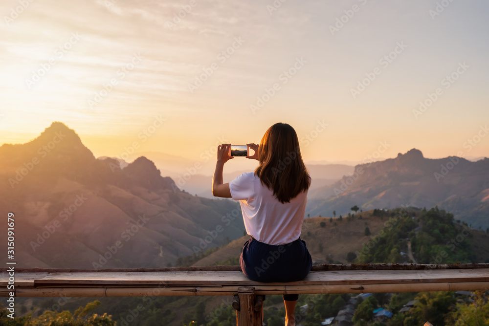 Young woman traveler taking photo with smart phone at sunset over the mountain - obrazy, fototapety, plakaty 