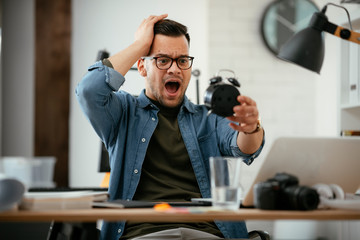 Young businessman getting late with his project. Handsome nervous man holding clock in office. 