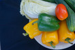 Fresh vegetables have yellow and green bell peppers, with turnips and cucumbers and tomatoes on a white plate with a black background.