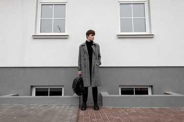 European urban young man in stylish autumn-winter outerwear with a fashionable leather black backpack in trendy boots stands on the street near a vintage wall. Handsome guy model in elegant clothes.