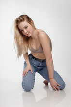 Portrait Of Sexy Caucasian Woman With Long Hair Posing In Beige Lingerie And Blue Jeans On White Studio Background. Model Tests Of Pretty Girl In Bra. Attractive Female Sitting On Floor On Her Knees