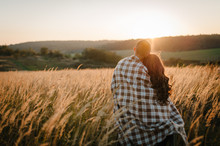 Young Couple Hugging, Standing Back, People Covered With Blanket, At Sunset In Autumn An Outdoor. At Field Grass On  Background Of Sun. Concept Of Friendly Family. Full Length. Close Up.