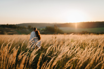 Couple hugging, people covered with blanket, at sunset in autumn an outdoor. at field grass on  background of sun.  full length standing back. Close Up. Place for text and design. selective focus.