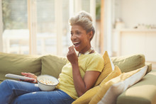 Mature Woman Eating Popcorn And Watching TV On Living Room Sofa