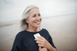 © Great Images/Caia Image - Smiling senior woman holding cell phone on winter beach