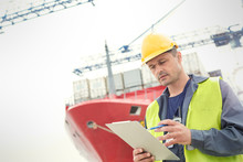 Dock Worker With Clipboard Below Container Ship At Shipyard