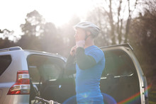 Male Cyclist Fastening Helmet At Back Of SUV In Sunny Parking Lot