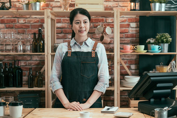 Wall Mural - Young female barista standing behind bar counter in cafe smiling and looking at customer while their order is ready on table. woman waitress in apron waiting for client to take cup of coffee.