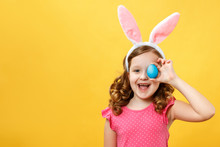 Happy Child In Bunny Ears Holds An Easter Blue Egg. Portrait Of A Little Girl On A Yellow Background