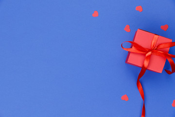 Blue background with red little hearts and a red box with a gift. Valentine's day background. On the right is a red gift box with a red ribbon. Close-up, free space on the left, horizontal. Concept of