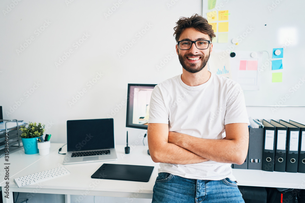 Portrait of confident graphic designer leaning on desk in office with arms crossed - obrazy, fototapety, plakaty 