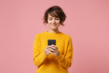Smiling Young Brunette Woman Girl In Yellow Sweater Posing Isolated On Pastel Pink Wall Background Studio Portait. People Lifestyle Concept. Mock Up Copy Space. Using Mobile Phone, Typing Sms Message.