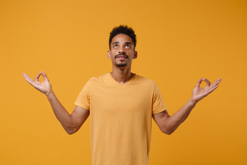 Young african american guy in casual t-shirt posing isolated on yellow orange background studio portrait. People lifestyle concept. Mock up copy space. Hold hands in yoga gesture, relaxing meditating.