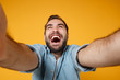 © ViDi Studio - Close up of laughing young man in casual blue shirt posing isolated on yellow orange wall background, studio portrait. People lifestyle concept. Mock up copy space. Doing selfie shot on mobile phone.