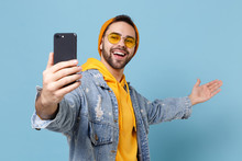 Smiling Young Hipster Guy In Fashion Jeans Denim Clothes Isolated On Pastel Blue Wall Background. People Lifestyle Concept. Mock Up Copy Space. Doing Selfie Shot On Mobile Phone Pointing Hand Aside.