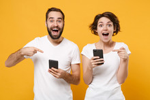 Surprised Couple Friends Bearded Guy Girl In White Blank Empty T-shirts Isolated On Yellow Orange Wall Background. People Lifestyle Concept. Mock Up Copy Space. Pointing Index Fingers On Mobile Phone.