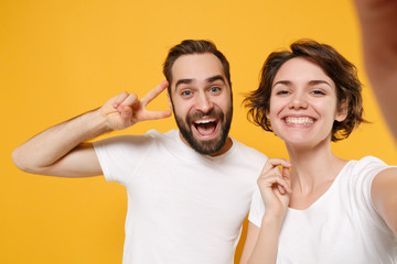 Wall Mural - Close up of couple friends bearded guy girl in white t-shirts isolated on yellow orange background. People lifestyle concept. Mock up copy space Doing selfie shot on mobile phone showing victory sign.