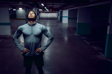 Wall Mural - Attractive tired caucasian sportsman in sportswear standing in garage with hands on hips and looking up.