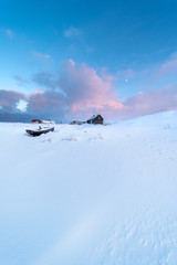 Wall Mural - wooden boat and wooden house on snow covered field in an arctic landscape at sunset with the moon in