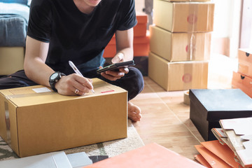 Thai young male online seller and start up small business owner using smartphone and writing address on cardboard box and packing parcels for delivery at home.