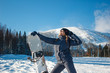 © Марина Демешко - Beautiful positive blond young woman in pigtails in grey costume is smiles, stands in the hero's pose and holds in hand snowboard on a background of ski resort. Winter sunny day