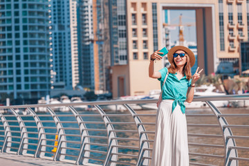 Wall Mural - Happy asian girl taking selfie photo on a smartphone while walking on a promenade in Dubai Marina district. Travel and lifestyle in United Arab Emirates