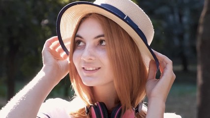 Wall Mural - Portrait of pretty positive teenage girl with red hair wearing straw hat and pink earphones smiling happily in camera.