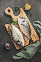 Fresh raw dorado fish, lemon and spices on a wooden cutting board, dark rustic background. Top view, flat lay, toned.